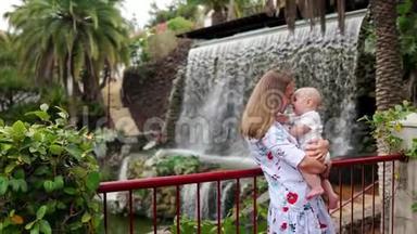 年轻的妈妈抱着一个孩子，和儿子一起玩，在公园的瀑布附近微笑和欢笑，同时去旅行